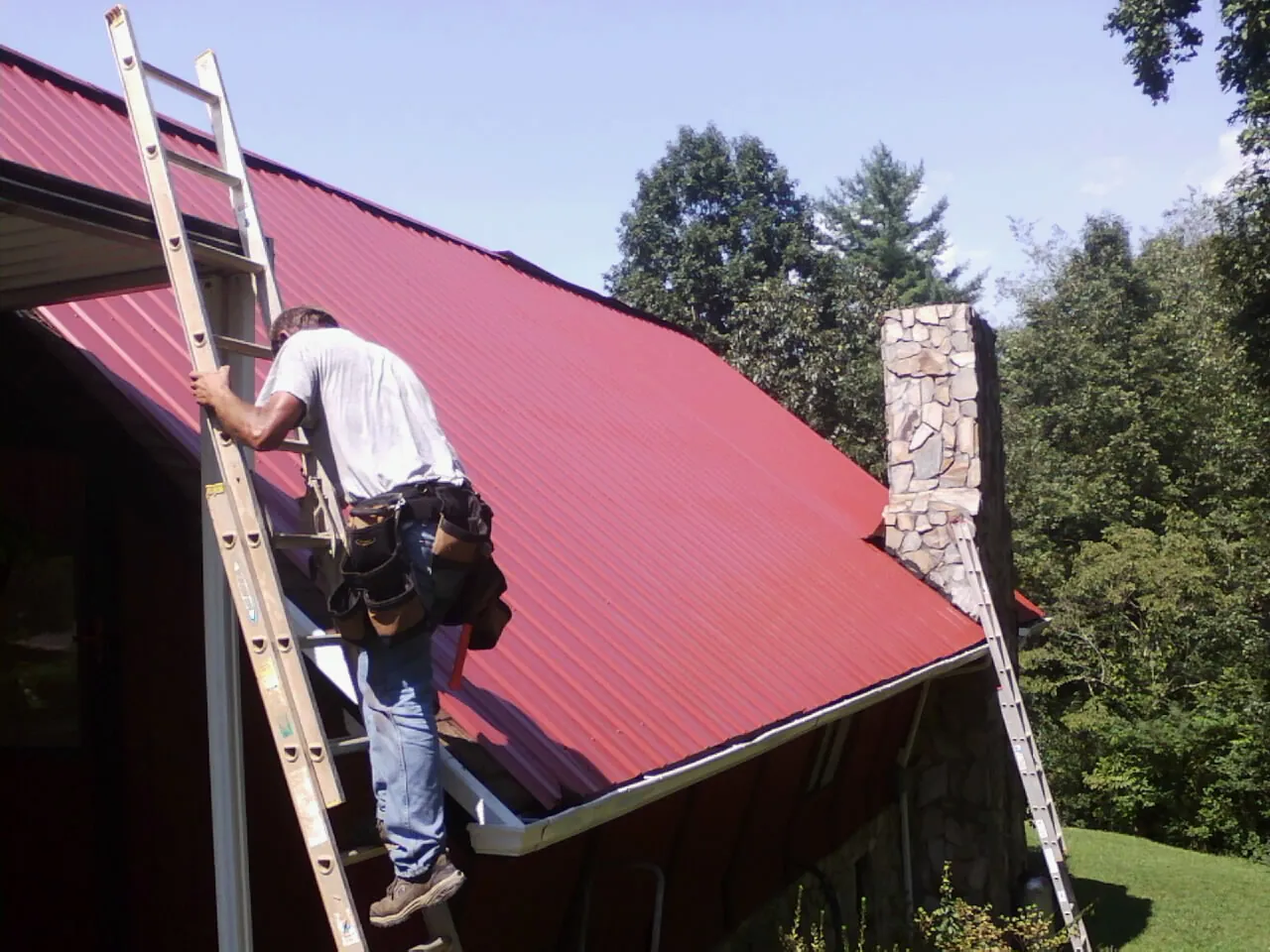 Roofer on a ladder beside a red metal roof and stone chimney — Soffit & Fascia Repair by Roofing Brothers in Artemas