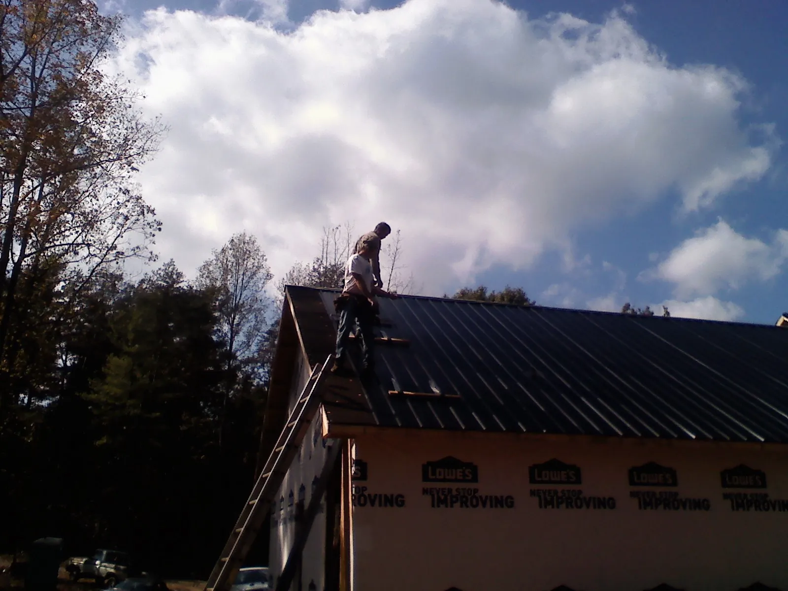 Roofer fastening dark metal panels high against a cloudy sky, from a Roof Inspection job in Artemas